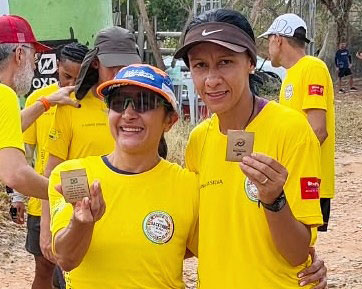 Lua, à esquerda, e Adriana posam abraçadas com camisetas amarelas do Brasil e seguram medalhas diante do pórtico da prova.